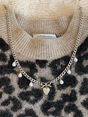 Silverfärgat halsband med pärlor och hjärta - Snyggt silverfärgat rostfritt halsband med kedja, små vita pärlor som hängande detaljer och ett hjärthänge med rosett i mitten. Perfekt accessoar för att lyfta din outfit med en söt och trendig touch.