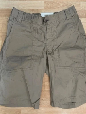Nya. Beige shorts från H2O med fickor - Säljer ett par beige shorts från H2O med stora framfickor och bakfickor med lock. Klassisk passform med bälteshällor och knappstängning. Perfekta för sommardagar och avslappnad stil.