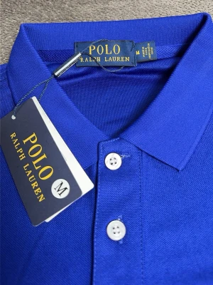 Blå polotopp från Polo Ralph Lauren - Snygg blå polotopp från Polo Ralph Lauren med klassisk krage och tre vita knappar framtill. T-shirten har korta ärmar och ikonisk broderad logga på bröstet. Perfekt för dig som vill ha en stilren och tidlös look.