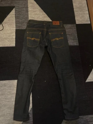 Mörkblå jeans från Nudie Jeans - Säljer ett par mörkblå jeans från Nudie Jeans i nyskick  med klassisk gul kontrastsöm och vågig detalj på bakfickan. Jeansen har rak passform och är tillverkade i kraftig denim. Perfekta för dig som gillar stilrena och tidlösa jeans. 29/32