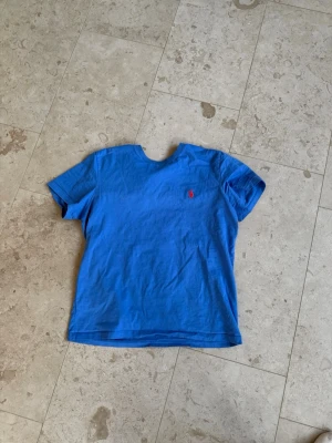 Blå t-shirt från Polo Ralph Lauren - Snygg blå t-shirt från Polo Ralph Lauren med klassisk röd logga broderad på bröstet. Modellen är custom slim fit och har korta ärmar. Perfekt för en avslappnad och stilren look.
