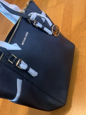 Michael Kors (Helt ny oanvänd) - Snygg svart handväska från Michael Kors i strukturerat läder med guldfärgade detaljer och logotyp. Väskan har dubbla handtag och dekorativ MK-berlock. Perfekt storlek för att få plats med allt du behöver och ger en lyxig känsla till din outfit. 100% äkta, köpt från USA.