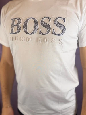 Vit Hugo Boss t-shirt med tryck - Säljer en vit t-shirt från Hugo Boss med stort BOSS-tryck i blått och vitt på bröstet. T-shirten har klassisk passform och korta ärmar, perfekt för dig som gillar stilrena märkeskläder. Materialet är mjukt och skönt bomull.