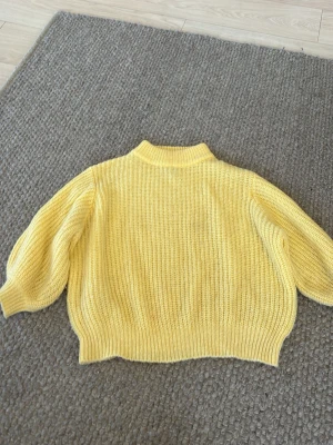 Gul stickad tröja med ballongärmar - Säljer en ljusgul stickad tröja med ballongärmar och ribbstickade muddar. Tröjan har en högre krage och är gjord i ett mjukt material som känns skönt mot huden. Perfekt för att addera lite färg till din outfit.