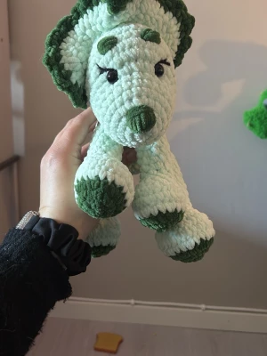 Virkad dinosaurie i grönt och vitt - Mysig handgjord dinosaurie i mjukt virkat garn. Färgerna är ljusgrön och mörkgrön med detaljer på tassar, nos och huvud. Den har stora ögon och en krage runt huvudet som påminner om en triceratops. Perfekt som dekoration eller gosedjur.