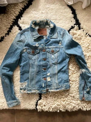 True religion  - Snygg ljusblå jeansjacka från true religion  med slitna detaljer och råa kanter. Klassisk modell med bröstfickor, knappar framtill och dragkedjor vid ärmsluten. Perfekt för dig som gillar en edgy streetstyle-look.