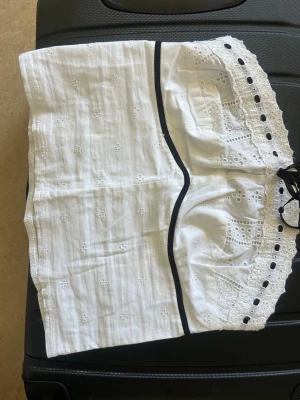 Vit broderad korsett med svarta detaljer - Vit korsett med broderade blommönster och svarta band längs kanterna. Framsidan har dekorativa hål och spetsdetaljer samt ett svart snörband för justering. Materialet är bomull med broderi och spets, perfekt för att skapa en elegant look.