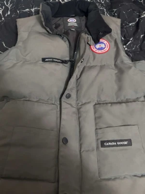 Grå dunväst från Canada Goose - Säljer en grå dunväst från Canada Goose med klassisk logotyp på bröstet och svarta detaljer vid kragen. Västen har två stora fickor framtill och dragkedja med tryckknappar. Perfekt för sommar .  dagar och snygg att kombinera med hoodie eller tröja. Priset kan diskuteras vid en snabb affär