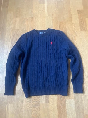 Mörkblå kabelstickad tröja Polo Ralph Lauren - Säljer en mörkblå kabelstickad tröja från Polo Ralph Lauren med klassisk rund hals och röd broderad logotyp på bröstet. Tröjan har långa ärmar och ribbade muddar vid ärmslut och nederkant. Perfekt för dig som gillar stilrena och tidlösa plagg.