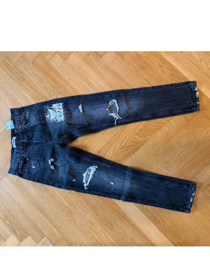Svarta slitna jeans med tryck - Coola svarta jeans med slitningar och tryckdetalj på ena benet. Raka ben , perfekt för en avslappnad streetstil. Jeansen har flera revor och en unik design som sticker ut.
