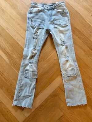 Ljusblå slitna jeans med raka ben - Säljer ett par ljusblå jeans med raka ben och flera stora slitningar och patchade detaljer framtill. Jeansen har råa, fransiga benslut och klassisk femficksdesign. Perfekta för en edgy streetstil.