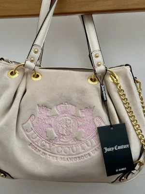 Juicy Couture scottie handväska  - Stilren beige handväska från Juicy Couture i mjuk mocka med ljusrosa broderad logga och emblem framtill. Väskan har vita handtag med guldfärgade detaljer och kedja, samt dragkedja upptill. Perfekt för dig som gillar en lyxig och trendig look.