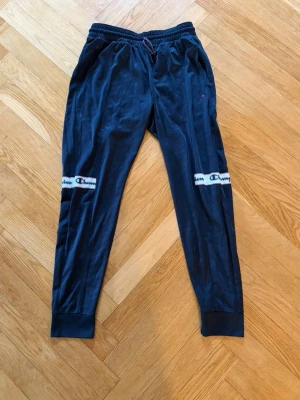 Svarta mjukisbyxor från Champion - Svarta mjukisbyxor från Champion med resår i midjan och dragsko. Byxorna har vita band med Champion-logga över knäna och muddar vid bensluten. Perfekta för en avslappnad stil.