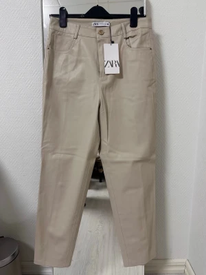 Beiga raka skinnbyxor från Zara - Snygga beiga byxor från Zara med rak passform och klassisk femficksdesign. Byxorna har hög midja, bälteshällor och stängs med knapp och dragkedja. Materialet är slätt och ger en stilren look, perfekt för dig som gillar minimalistisk stil.