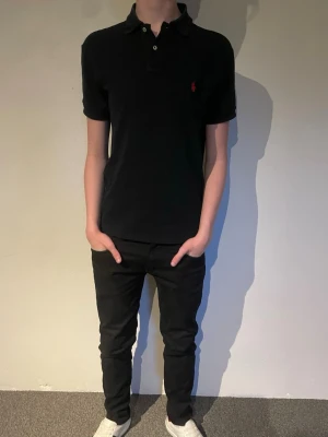 Svart pikétröja från Ralph Lauren - Klassisk svart pikétröja från Ralph Lauren med korta ärmar och liten röd logga på bröstet. Tröjan har krage och knäppning med två knappar framtill. Perfekt till jeans eller shorts för en stilren look.