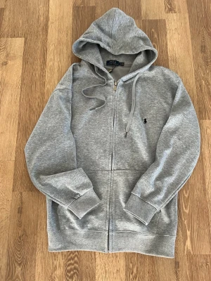 Grå zip-hoodie från Polo Ralph Lauren - Snygg grå hoodie med dragkedja från Polo Ralph Lauren. Klassisk design med huva, dragsnören och broderad svart logga på bröstet. Mjuk bomullsblandning och ribbade muddar vid ärmslut och nederkant. Perfekt för en chill och stilren look.