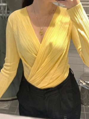 Gul omlottbody Ginatricot  - Snygg gul body med omlott-design och djup V-ringning. Långa ärmar och plisserat tyg ger en trendig look. Perfekt att styla med både jeans och kjol. Materialet är stretchigt och bekvämt. Stängs med tryckknappar i grenen.