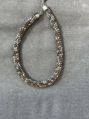 Flätat halsband med pärlor i metallic - Säljer ett flätat halsband med små pärlor i silver, brons och guldtoner. Halsbandet har en rund form och stängs med en metallkedja och lås. Perfekt för dig som gillar unika accessoarer med coola färgkombinationer.
