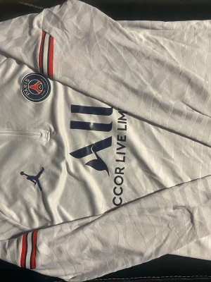 Vit PSG långärmad träningströja - Säljer en vit långärmad träningströja från PSG x Jordan med subtilt mönster i tyget. Tröjan har dragkedja vid halsen, PSG-logga på bröstet och röda samt svarta detaljer på ärmarna. Perfekt för dig som gillar fotboll och vill ha en snygg sportlook.