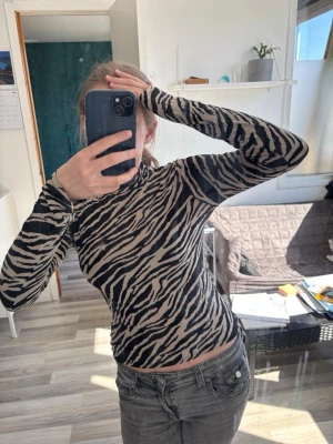 Zebramönstrad polotopp i beige och svart - Säljer en zebramönstrad polotopp i beige och svart. Toppen är tight och har långa ärmar samt hög krage. Perfekt för dig som vill sticka ut med ett djurmönster. Materialet känns mjukt och stretchigt, troligen polyester eller bomullsmix.