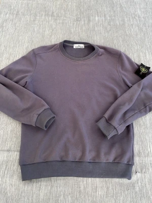 Blå sweatshirt från Stone Island - Snygg blå sweatshirt från Stone Island med klassisk rund halsringning och ribbade muddar. Tröjan har den ikoniska Stone Island-loggan på vänster ärm och är tillverkad i mjuk bomull för maximal komfort.