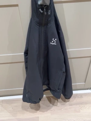 Svart Haglöfs Kaise GTX regnjacka - Snygg svart regnjacka från Haglöfs, modell Kaise GTX. Jackan har huva, hel dragkedja och logga på bröstet. Tillverkad i slitstarkt och vattentätt material, perfekt för regniga dagar. Enkel och stilren design som funkar till det mesta. Använd 3 gånger mer eller mindre ny!!! ,Storlek S men du kan skjustera  den till din anpassning.