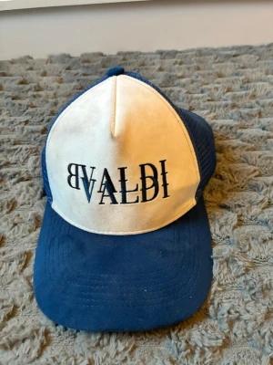 Blå och vit keps från Bvaldi - Snygg keps från Bvaldi i blått och vitt med broderad logga framtill. Kepsen har klassisk trucker-modell med mesh på sidorna och justerbar snapback baktill. Perfekt för dig som vill ha en stilren accessoar med streetwear-vibe.