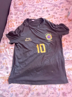 Svart Brasilien fotbollströja #10 - Svart fotbollströja med Brasilien-emblem och nummer 10 i gult framtill. Tröjan har rund hals och korta ärmar, samt texten 'Joga Bonito' tryckt på bröstet. Perfekt för dig som älskar fotboll och vill representera Brasilien.