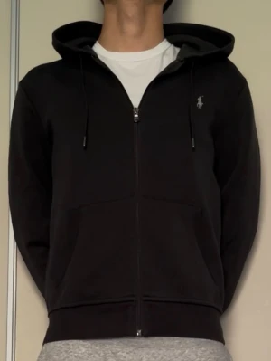 Svart hoodie från Polo Ralph Lauren - Svart zip-hoodie från Polo Ralph Lauren med huva. Klassisk logga broderad på bröstet i vitt. Tidlös design som passar med flera outfits!                        Hör gärna av er vid frågor🙌