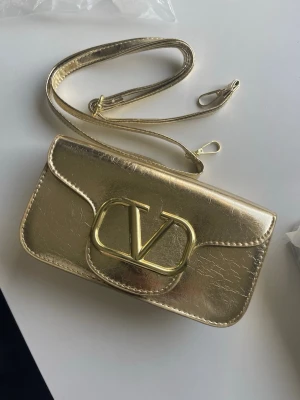 Guldig axelväska i metallic, tar ej bud - Snygg axelväska i metallic guld med slät, glansig yta och rektangulär form. Väskan har en lång axelrem i samma material och färg. Perfekt för dig som vill sticka ut med en trendig accessoar. Märke och exakt material är okänt.