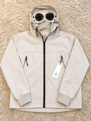 Beige vindjacka med huva från CP Company  - Stilren beige vindjacka med svart dragkedja och två snedställda fickor med dragkedja. Jackan har justerbar huva med snörning och ribbade muddar vid ärmsluten. Perfekt för blåsiga dagar och enkel att matcha med olika outfits.