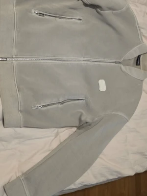 Emporio Armani Beige ziptröja och matchande Byxa - Snygg ljusgrå ziptröja från Emporio Armani med två dragkedjeförsedda fickor och broderad logga på bröstet strlk Large. Tröjan har en stilren design och är tillverkad i mjukt bomullsmaterial. Perfekt för en clean och modern look. Matchande byxa i samma färg och strlk Large