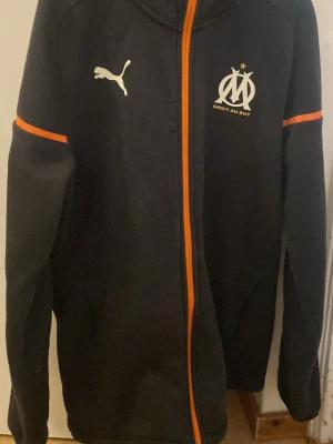 Svart Marseille hoodie från Puma - Svart hoodie med dragkedja från Puma och Marseille-logga på bröstet. Orange detaljer längs dragkedjan och ärmarna. Tillverkad i mjukt material och har huva. Perfekt för dig som gillar fotboll och vill ha en sportig look. Köpt från Zalando för 1200 kr 