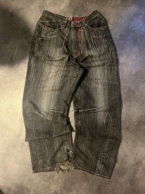 Gråa AKDMKS jeans med patchar - Unika grå jeans från AKDMKS med vertikala ränder, röda detaljer och flera färgglada patchar på bakfickan. Vida ben och slitna detaljer nertill ger en cool streetwear-vibe. Perfekta för dig som gillar statement-plagg.