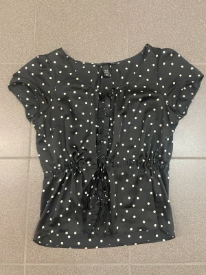 Svart prickig blus från H&M - Svart blus från H&M med vita prickar, korta ärmar och knytband i midjan. Söt volangdetalj längs knäppningen framtill. Perfekt för en trendig och lekfull look. Tillverkad i ett lätt och luftigt material. Pris kan sänkas vid snabbt köp💕💕👍🏼