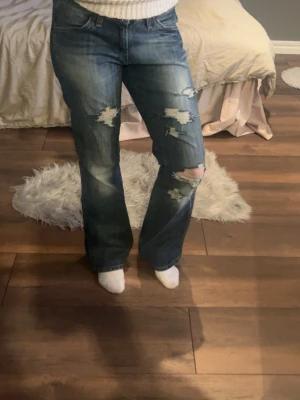 Blå slitna bootcut jeans - Säljer ett par blå bootcut jeans med slitningar och hål på framsidan. 