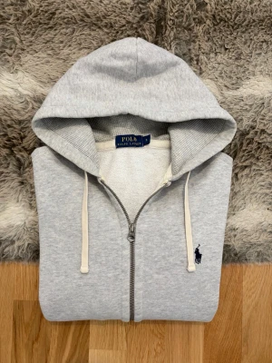 Ljusgrå Polo Ralph Lauren Hoodie - Hej! 👋 Säljer en riktigt stilren och tidlös ljusgrå Polo Ralph Lauren zip-up i storlek S. Den har den klassiska marinblå pony-loggan broderad på bröstet 🐎 Tröjan är i mycket gott skick, välvårdad och fräsch. 100% äkta med tvättlappar kvar (ingen QR-kod på denna modell). En zip-up är det ultimata basplagget som funkar till allt och är otroligt smidig att bära. Fraktar snabbt! 🚛📦💬💯 
