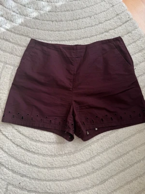 Vinröda shorts med broderade detaljer - Snygga vinröda shorts med broderade håldetaljer längst ner på benen. Modellen är högmidjad och har fickor i sidorna. Perfekta för sommaren och tillverkade i ett mjukt bomullstyg.