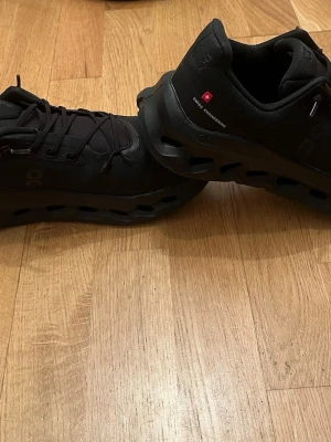 Svarta On Cloud sneakers - Snygga helsvarta sneakers från On med modern design och unika hål i sulan. Skorna har diskret logga på sidan, svarta skosnören och detaljer med texten 'Swiss Engineering'. Ovandelen är i textil och sulan i gummi för extra komfort.
