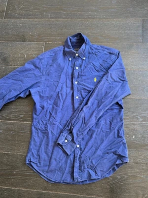 Blå skjorta från Ralph Lauren - Klassisk blå skjorta från Ralph Lauren med gul broderad logga på bröstet. Skjortan har lång ärm, knappar framtill och button-down krage. Perfekt för en stilren look.