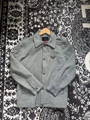 Grå overshirt från Prada - Snygg grå overshirt från Prada med klassisk krage och knäppning framtill. Bröstficka med ikonisk Prada-logga i svart på vänster sida. Rak passform och diskreta sömmar ger en clean look. Perfekt för lager-på-lager-stil.