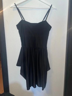 Svart asymmetrisk klänning - Svart kortklänning med smala axelband och asymmetrisk nederkant. Klänningen har en enkel, stilren design och är tillverkad i ett mjukt tyg som ger ett snyggt fall. Perfekt för dig som gillar minimalistisk stil.