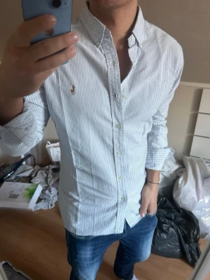 Randig skjorta från Ralph Lauren - Snygg vit skjorta med tunna blå ränder från Ralph Lauren. Klassisk button-down krage, broderad logga på bröstet och långärmad modell. Skjortan har en slim passform och är tillverkad i bomull. Perfekt för en stilren look. (små defekter på bild 4 och 5)
