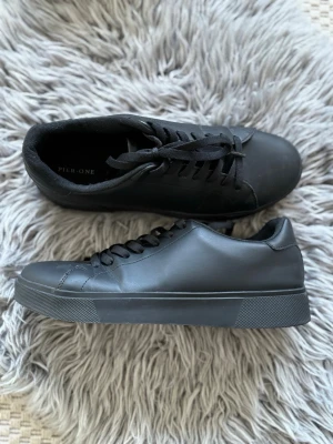 Svarta sneakers - Stilrena svarta sneakers i slät syntet med matchande svarta skosnören och sula. Klassisk låg modell med enkel design som passar till det mesta. Perfekta för dig som gillar minimalistisk stil. Oanvända!