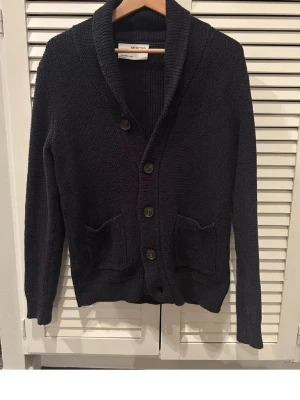 Mörkblå stickad kofta med sjalkrage - Mysig mörkblå stickad kofta från Selected med sjalkrage och fyra knappar framtill. Två praktiska fickor på framsidan och ribbade muddar vid ärmslut och nederkant. Perfekt för lager på lager och en avslappnad stil.