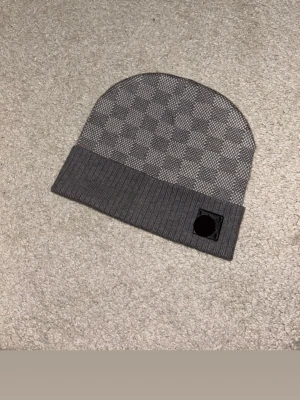 Grå rutmönstrad mössa med patch - Snygg grå mössa med diskret rutmönster och bred ribbad kant. På kanten sitter en svart patch i läderimitation som ger en exklusiv touch. Perfekt accessoar för kalla dagar och passar till de flesta outfits. Fick den i present har bara använt den 1 gång.