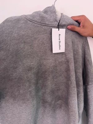Grå hoodie från Acne Studios - Snygg grå hoodie från Acne Studios med tryck 'Acne Studios STOCKHOLM 1996' på framsidan. Klassisk passform med mjukt material och ribbade muddar. Perfekt för en chill och stilren look.