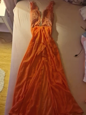 Orange långklänning från Ginger Fizz - Snygg orange långklänning från Ginger Fizz med spetsdetaljer upptill och djup v-ringning. Klänningen har ärmlös design och ett lätt, flödande tyg som ger en elegant look. Perfekt för speciella tillfällen när du vill sticka ut. 