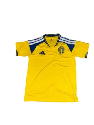 Svensk landslagströja Adidas gul - Gul fotbollströja från Adidas med Sveriges landslagsemblem på bröstet. Tröjan har blå detaljer, vita ränder på axlarna och en blå V-ringad krage. Tillverkad i lätt och ventilerande material, perfekt för träning eller match.