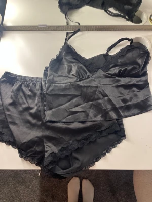 Svart satinset med spetsdetaljer - Snyggt svart set i satin med linne och shorts. Linnet har tunna justerbara axelband och spetskant vid urringningen. Shortsen har elastisk midja och spetskant nedtill. Perfekt för en lyxig känsla hemma. Materialet är glansigt och mjukt mot huden.
